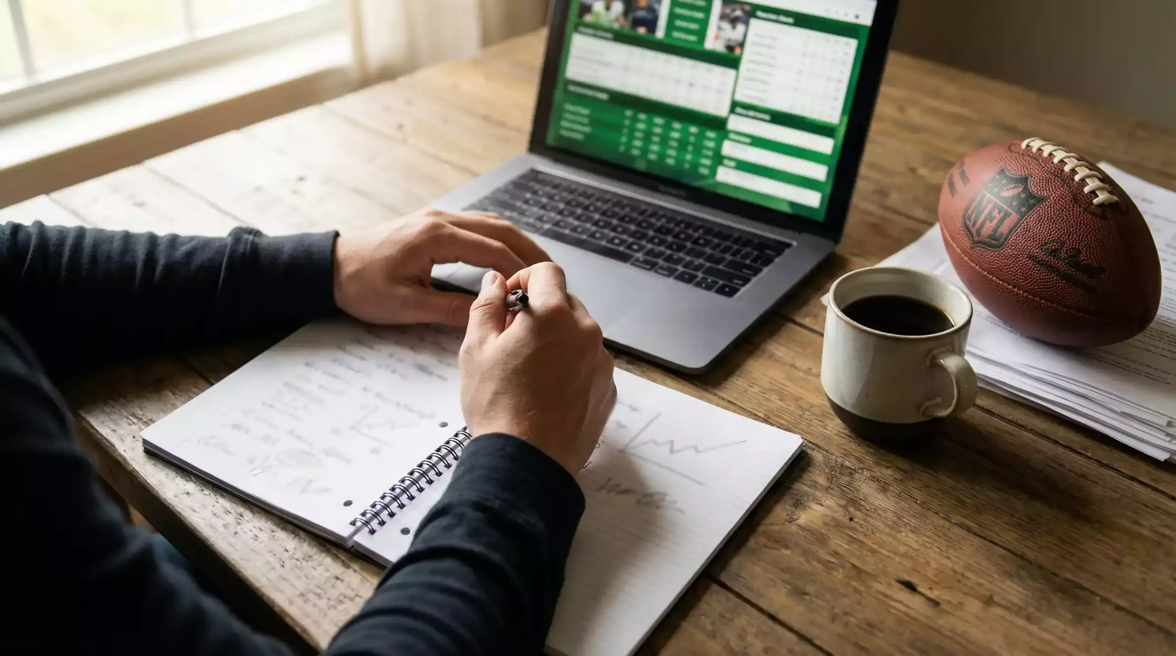 Persona analizando estadísticas de fútbol americano con libreta y portátil en una mesa